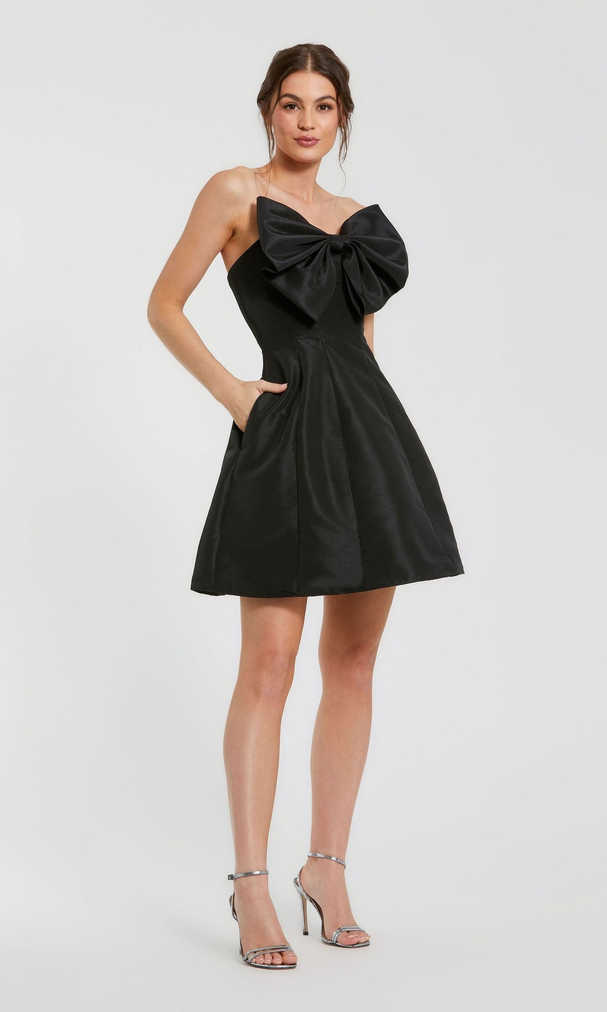 A woman models the Mac Duggal 49210 Strapless Short Bow Party Dress with a large chest bow, hands in pockets, and silver heels, posing against a white background.